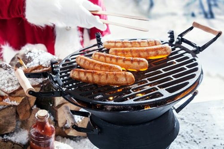 Ein Weihnachtsmann grillt Würstchen auf einem runden Grill im Schnee.